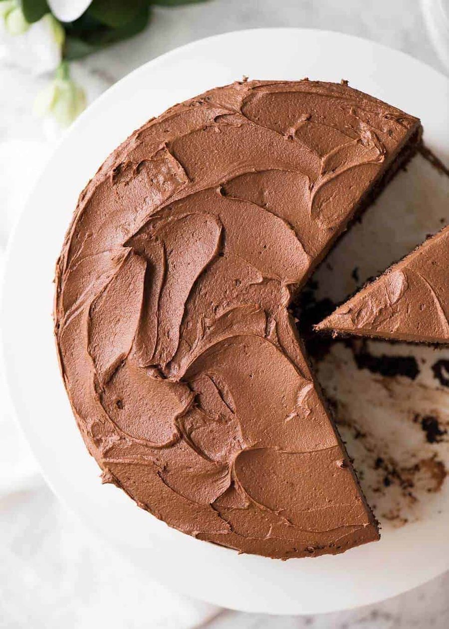 Chocolate Cake with Chocolate Buttercream Frosting on a white cake platter with a piece cut out, photographed from overhead.