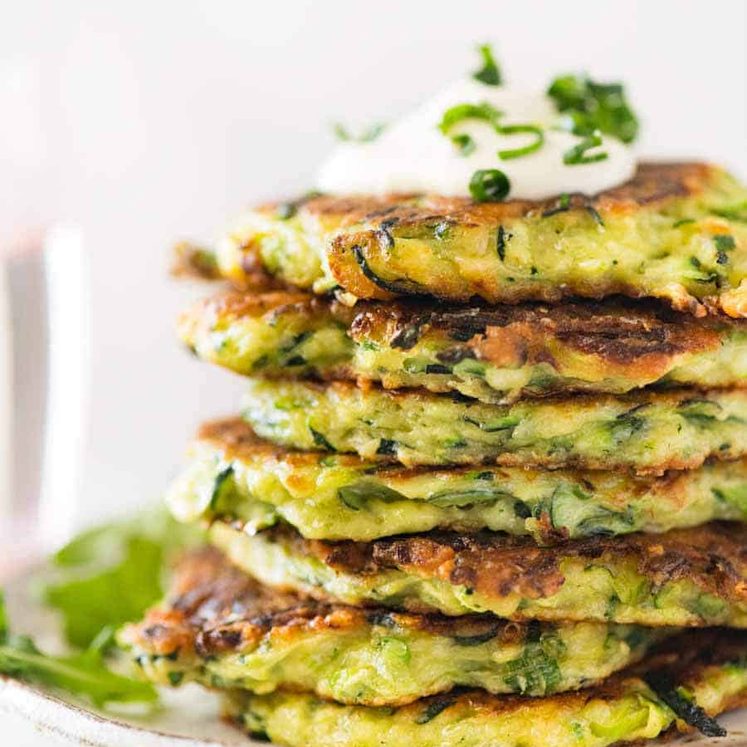 Stack of Crispy Zucchini Fritters with a dollop of sour cream on a rustic brown plate with a side of arugula salad,