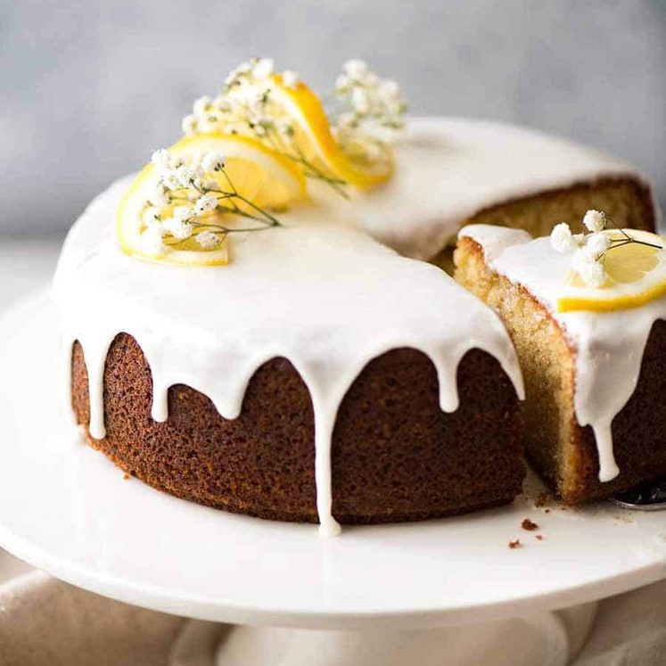 Lemon Cake with drippy lemon glaze decorated with lemon slices and flowers, on a white cake platter with a piece cut out