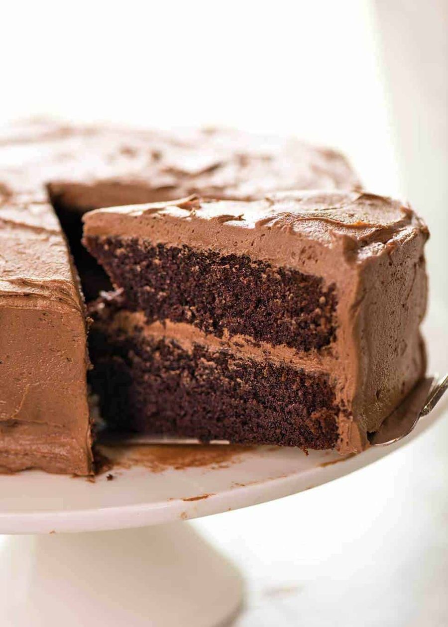 A photo showing half a Chocolate Cake frosted with Chocolate Buttercream on a white cake stand with a large slice partially pulled out.